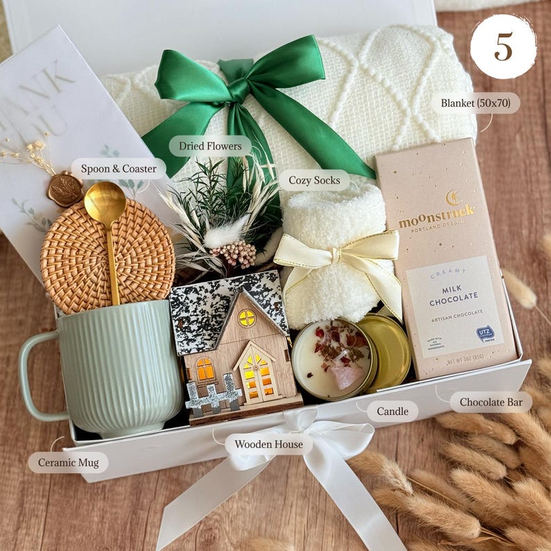 May include: A gift box containing a white knit blanket with a green ribbon, a light green ceramic mug, a rattan coaster, a gold spoon, dried flowers, white cozy socks, a wooden house ornament, a candle, and a Moonstruck milk chocolate bar.