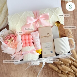 May include: A gift box filled with self-care items, including a white blanket, a pink hand lotion, a pink candle, a white ceramic mug, a chocolate bar, a bouquet of dried flowers, cozy socks, and a spoon and coaster.