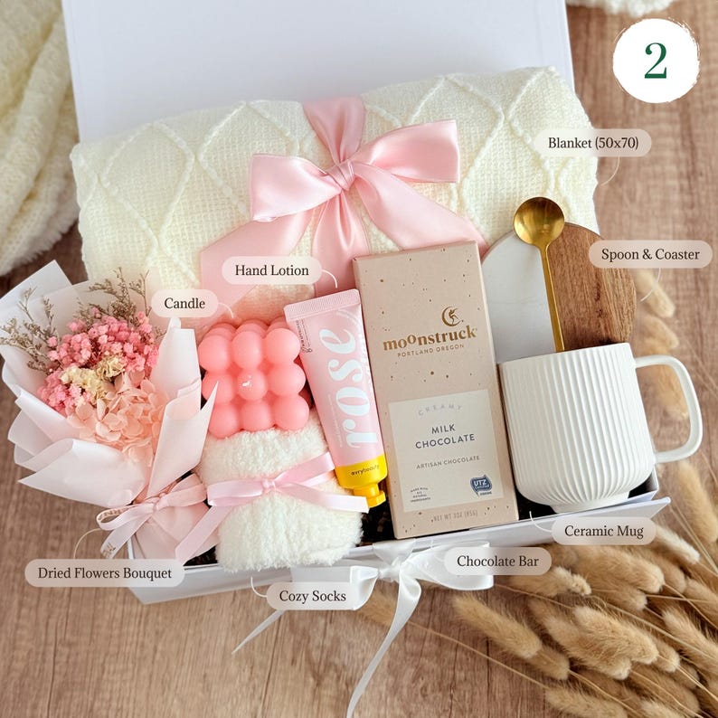 May include: A gift box filled with self-care items, including a white blanket, a pink hand lotion, a pink candle, a white ceramic mug, a chocolate bar, a bouquet of dried flowers, cozy socks, and a spoon and coaster.