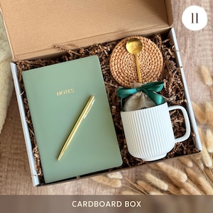 Peut inclure: Coffret cadeau contenant un carnet vert sauge avec "NOTES" en lettres dor&eacute;es, un stylo dor&eacute;, une tasse blanche c&ocirc;tel&eacute;e, une cuill&egrave;re dor&eacute;e, un dessous de verre tiss&eacute; et un petit sac en toile de jute nou&eacute; avec un ruban vert. La bo&icirc;te est &eacute;tiquet&eacute;e "CARDBOARD BOX".