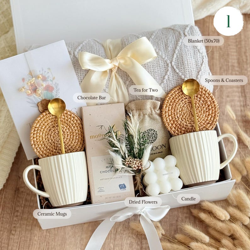 May include: A white gift box filled with cozy items for a relaxing evening. The box includes a white blanket, two ceramic mugs, a chocolate bar, tea, a candle, dried flowers, and two woven coasters with gold spoons.
