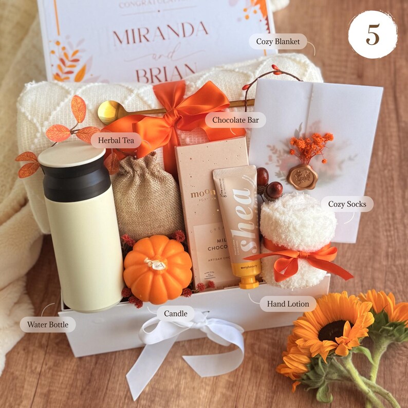 May include: A gift box filled with a variety of items, including a water bottle, candle, chocolate bar, hand lotion, and cozy socks. The box is decorated with orange ribbons and fall-themed accents, such as sunflowers and a pumpkin-shaped candle.