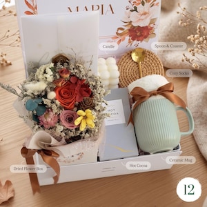 May include: A gift box containing a dried flower bouquet, a white bubble candle, a rattan coaster, a gold spoon, a packet of hot cocoa, and a ribbed light green ceramic mug wrapped with a brown ribbon. The box features floral graphics and the name MARIA.