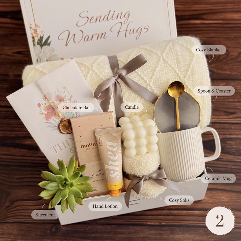 May include: A gift box containing a cream-colored knit blanket with a grey ribbon, a chocolate bar, a tube of shea hand lotion, a bubble candle, a pair of fuzzy socks, a succulent, and a ribbed ceramic mug with a gold spoon on a grey coaster.