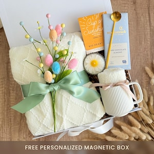 May include: A white gift box filled with a white blanket, a mug, a candle, a tea bag, a chocolate bar, a spoon, and a sprig of pastel colored Easter eggs. The box is decorated with a green ribbon and the text "FREE PERSONALIZED MAGNETIC BOX".