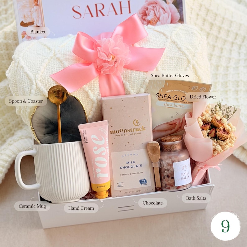 May include: A gift set containing a white cable-knit blanket with a pink bow, a white ribbed ceramic mug, a gold spoon, a gray coaster, a tube of rose hand cream, a Moonstruck milk chocolate bar, Shea-Glo gloves, bath salts, and a dried flower bouquet.