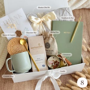 May include: A gift box containing a grey blanket, a sage green notebook with a gold pen, a Moonstruck milk chocolate bar, a ceramic mug, a rattan coaster, a gold spoon, a small burlap bag of herbal tea, a candle, and cinnamon sticks.