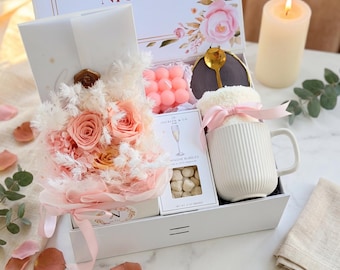 May include: A white gift box with a floral arrangement, mug, candle, and other items. The box is decorated with pink roses and the name "Nicole". The mug is white with vertical lines and a pink bow.