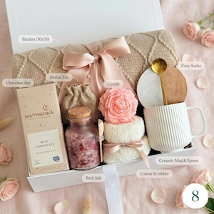 May include: A self-care gift box containing a beige blanket (approx. 127x178cm), a chocolate bar, herbal tea, a pink rose candle, bath salts, a white mug with a gold spoon, and cosy socks. The box is tied with a pink ribbon.