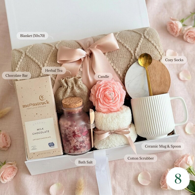May include: A self-care gift box containing a beige blanket (approx. 127x178cm), a chocolate bar, herbal tea, a pink rose candle, bath salts, a white mug with a gold spoon, and cosy socks. The box is tied with a pink ribbon.
