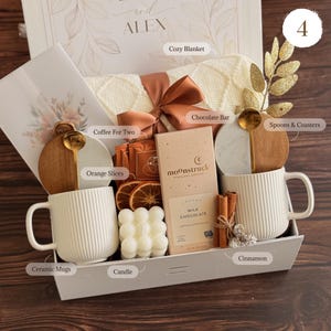 May include: A gift box containing two white ribbed ceramic mugs, a cream-colored knit blanket with a copper ribbon, a Moonstruck milk chocolate bar, a bubble candle, dried orange slices, cinnamon sticks, gold spoons, and marble-wood coasters.