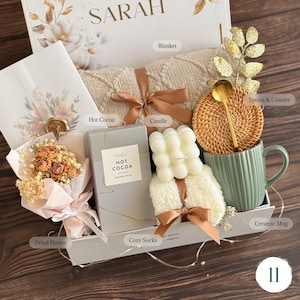 May include: A gift box containing a beige textured blanket tied with a brown ribbon, a box of hot cocoa, a white bubble candle, a pair of white fuzzy socks, a sage green ceramic mug, a rattan coaster, a gold spoon, and a dried flower bouquet.