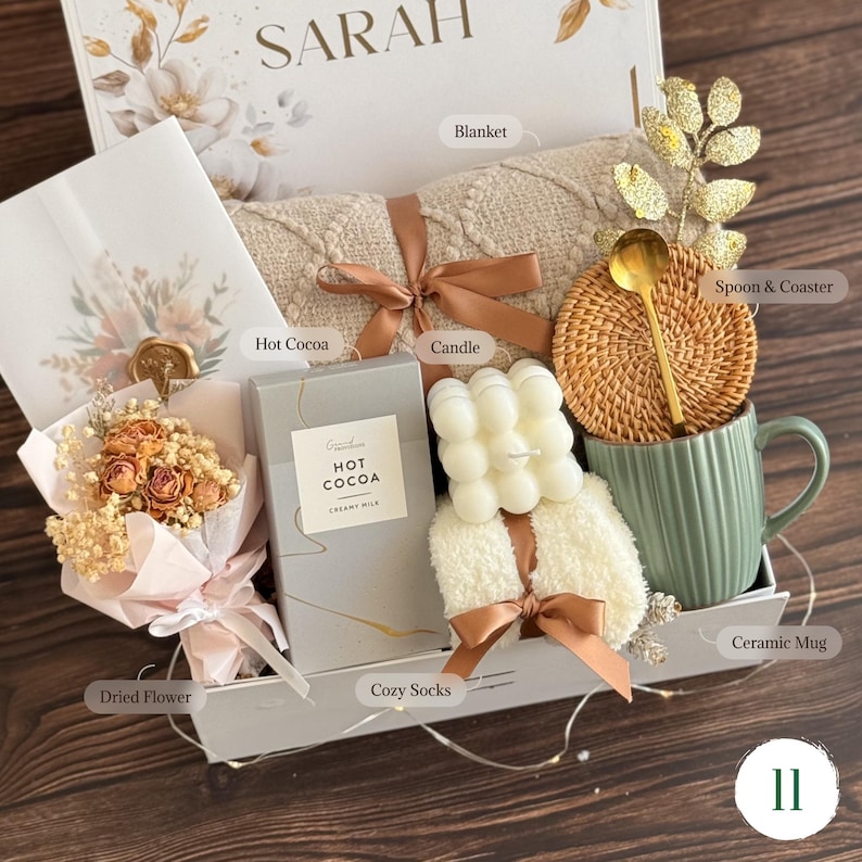 May include: A gift box containing a beige textured blanket tied with a brown ribbon, a box of hot cocoa, a white bubble candle, a pair of white fuzzy socks, a sage green ceramic mug, a rattan coaster, a gold spoon, and a dried flower bouquet.