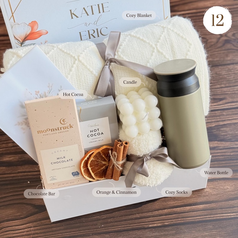 May include: A gift set containing a white knit blanket, a Moonstruck milk chocolate bar, a box of hot cocoa, a white bubble candle, a pair of fuzzy socks, dried orange slices with cinnamon sticks, and an olive green water bottle with a black lid.