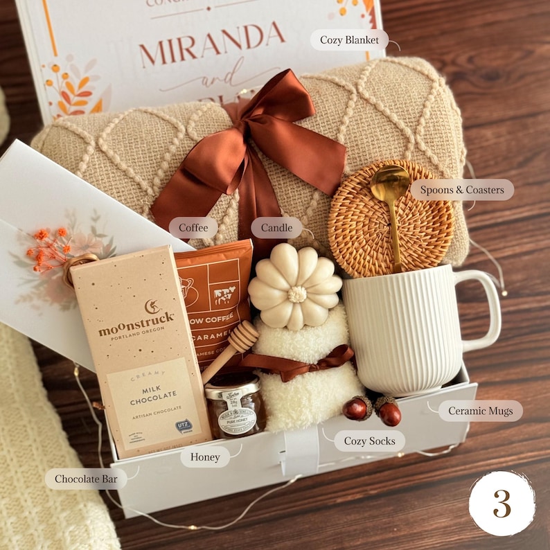 May include: A gift box containing a beige knit blanket with a brown ribbon, a Moonstruck milk chocolate bar, a bag of coffee, a flower-shaped candle, a jar of honey, a wooden honey dipper, a white ribbed ceramic mug, and a rattan coaster with a gold spoon.