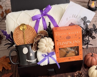 May include: A gift box containing a white cable-knit blanket tied with a purple ribbon, a black skull mug with a gold spoon, a round woven coaster, dried citrus slices, a white pumpkin candle, and a box of Rustic Bakery pumpkin cookies.