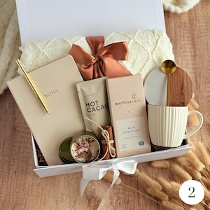 May include: A gift box filled with a cream-colored blanket, notebook, pen, hot cocoa, milk chocolate, a candle, a mug, and a wooden spoon. The box is tied with a brown ribbon and a white bow. The notebook reads "NOTES".