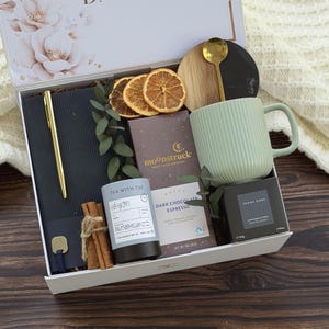May include: A gift box containing a black notebook with a gold pen, a light green ribbed mug, a gold spoon, a Moonstruck dark chocolate bar, a Tea With Tae tin, a small Aroma Home candle, dried orange slices, and cinnamon sticks.