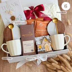 May include: A gift box containing two white ribbed ceramic mugs with gold spoons, two round wood and marble coasters, a Moonstruck dark chocolate espresso bar, two Copper Cow salted caramel coffee packets, a wooden house, and a white blanket.
