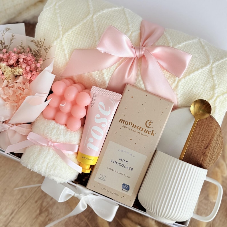 May include: A gift box containing a cream-coloured blanket, pink ribbon, dried flowers, a pink bubble candle, rose-scented lotion, a milk chocolate bar, a white mug, and a wooden spoon. The box is tied with a white ribbon.
