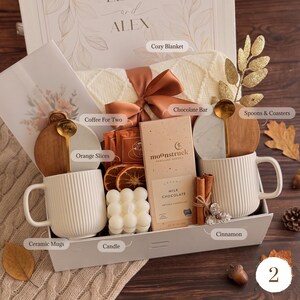 May include: A gift box containing two white ribbed ceramic mugs, a Moonstruck milk chocolate bar, a white bubble candle, dried orange slices, cinnamon sticks, two gold spoons, two marble and wood coasters, and a cream-colored knit blanket.