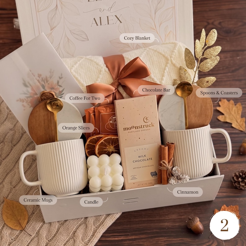 May include: A gift box containing two white ribbed ceramic mugs, a Moonstruck milk chocolate bar, a white bubble candle, dried orange slices, cinnamon sticks, two gold spoons, two marble and wood coasters, and a cream-colored knit blanket.