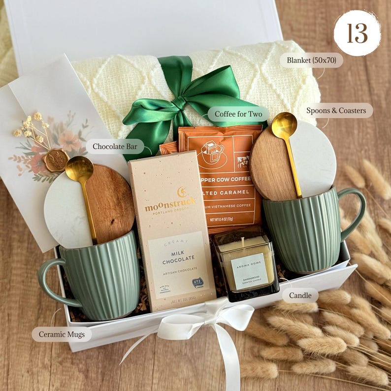 May include: A gift box containing a cream-colored knit blanket, two sage green ribbed ceramic mugs, two gold-tone spoons, two round coasters, a Moonstruck milk chocolate bar, a Copper Cow Coffee packet, and a small square candle in a glass jar.