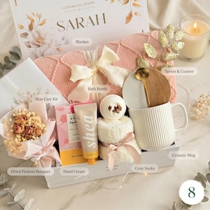 May include: A gift box with a variety of items, including a pink blanket, bath bomb, skincare kit, dried flowers, hand cream, cozy socks, a ceramic mug, and a spoon with a coaster. The box is decorated with a bow and the word "SARAH".