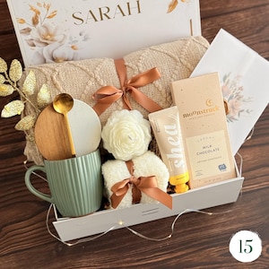 May include: A gift box containing a beige cable knit blanket with a brown ribbon, a sage green ribbed mug with a wooden lid and gold spoon, a white flower-shaped candle, a tube of shea hand cream, and a Moonstruck milk chocolate bar.