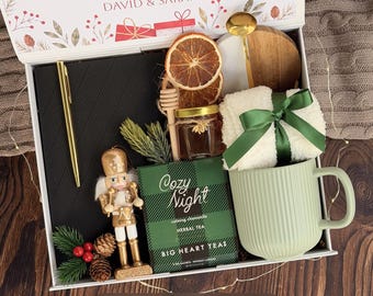May include: A gift box labelled DAVID & SARAH containing a black notebook, gold pen, nutcracker ornament, dried orange slices, honey jar with dipper, gold spoon, white fuzzy socks with a green ribbon, and a green Cozy Night tea box.