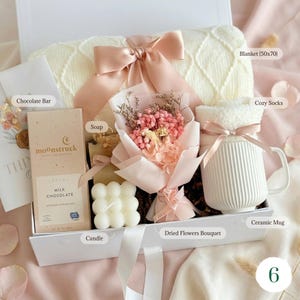 May include: A gift box filled with a cream-colored blanket (50x70 inches), cozy socks, a ceramic mug, a chocolate bar, soap, a candle, and a dried flower bouquet. The box is tied with a pink ribbon.