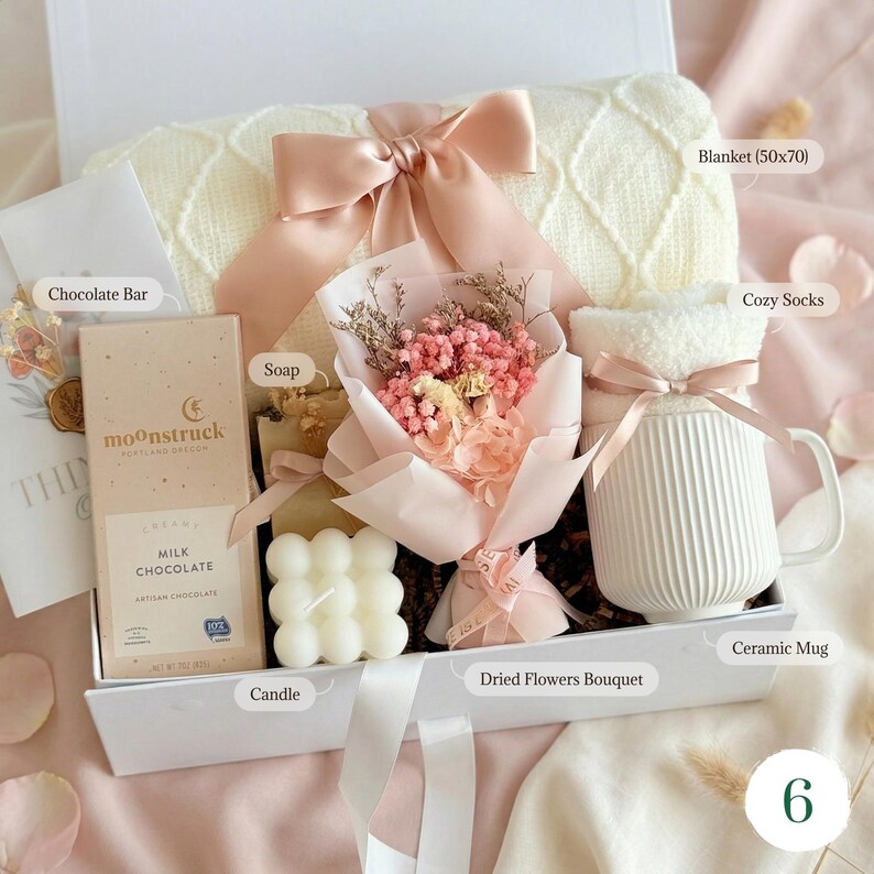 May include: A gift box filled with a cream-colored blanket (50x70 inches), cozy socks, a ceramic mug, a chocolate bar, soap, a candle, and a dried flower bouquet. The box is tied with a pink ribbon.