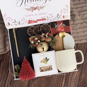 May include: A gift box containing a black notebook, a gold pen, a red honeycomb tree candle, a box of Jocelyn & Co roasted pistachios, a white ribbed mug, a gold spoon, a wooden coaster, and a decorative jingle bell with dried fruit.