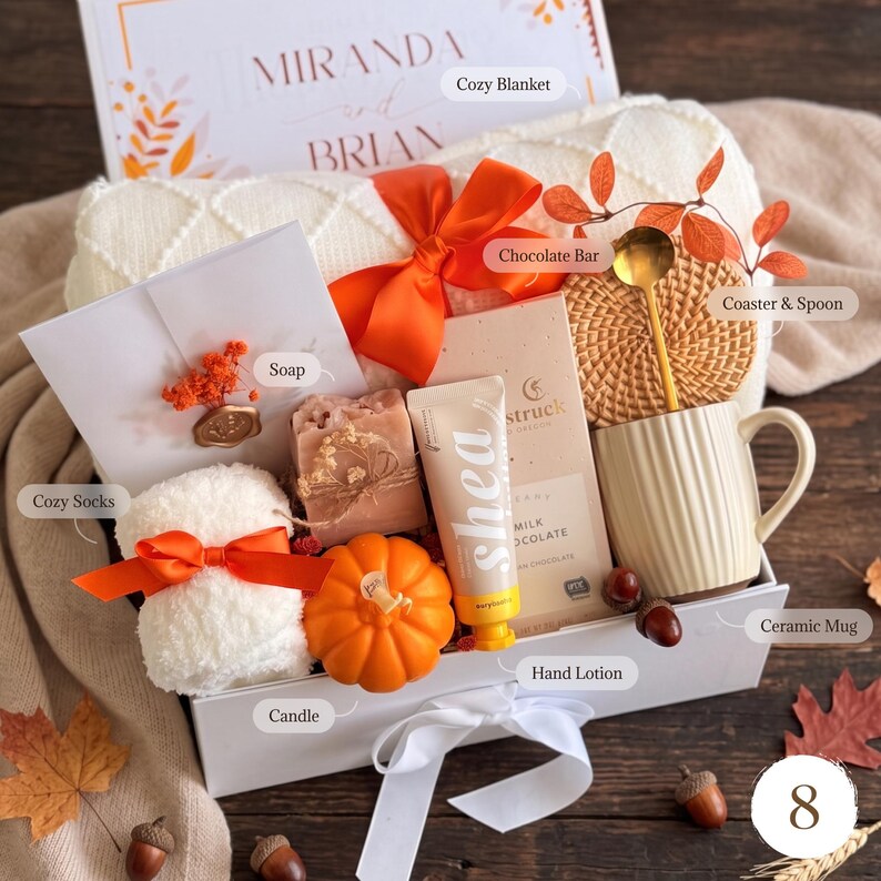 May include: A gift box containing a white knit blanket, a pair of white fuzzy socks with an orange ribbon, a bar of soap, a pumpkin-shaped candle, a tube of shea hand lotion, a chocolate bar, a round coaster, a gold spoon, and a ceramic mug.
