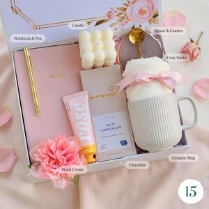 May include: A gift box with a pink theme, containing a pink notebook and pen, a candle, a bar of chocolate, hand cream, a ceramic mug with cozy socks, and a spoon on a coaster. The box is decorated with floral accents.