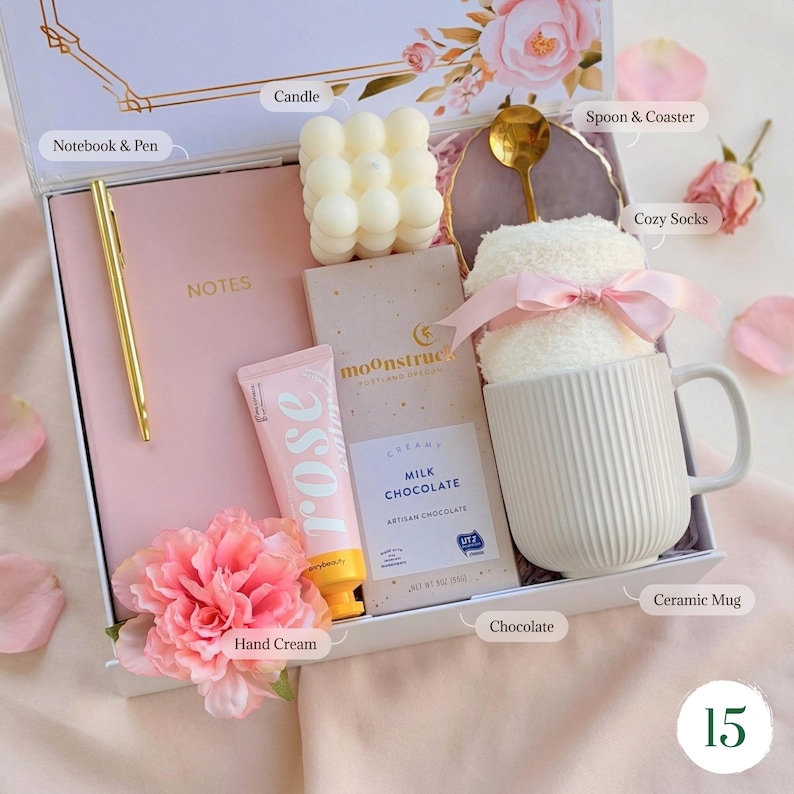 May include: A gift box with a pink theme, containing a pink notebook and pen, a candle, a bar of chocolate, hand cream, a ceramic mug with cozy socks, and a spoon on a coaster. The box is decorated with floral accents.