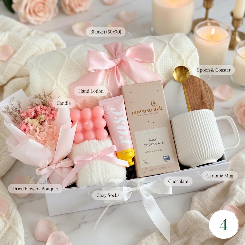 May include: A gift box with a white blanket (127x178 cm), pink bow, dried flower bouquet, pink candle, hand lotion, cozy socks, chocolate bar, ceramic mug, and a spoon with a wooden coaster. The items are arranged on a white surface.