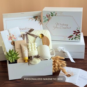 May include: A white gift box with a variety of items, including a mug, candle, and skincare products. The box is decorated with floral accents and the text "Wishing You Comfort". A smaller box is visible with the text "Sending".