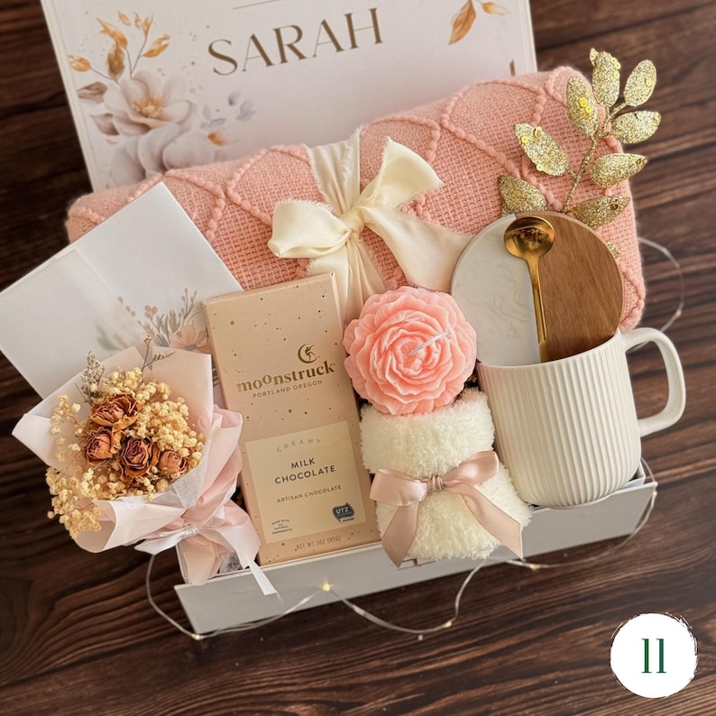 May include: A gift box containing a pink knit blanket with a cream ribbon, a Moonstruck milk chocolate bar, a pink rose-shaped candle, a white ribbed mug with a marble and wood lid, a gold spoon, a small dried flower bouquet, and a card labeled SARAH.