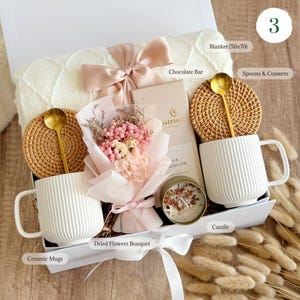 May include: A gift box containing a white knit blanket, two white ribbed ceramic mugs, two round woven coasters, two gold-toned spoons, a chocolate bar, a small candle, and a bouquet of pink and white dried flowers wrapped in light pink paper.