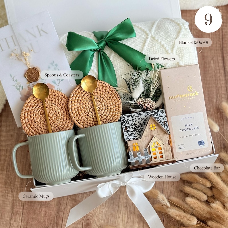 May include: A gift box containing two ribbed sage green ceramic mugs, two round woven rattan coasters, two gold-toned spoons, a white knit blanket with a green ribbon, a small wooden house, dried flowers, and a Moonstruck milk chocolate bar.
