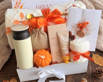 May include: A gift box containing a cream-colored tumbler, a burlap pouch, an orange pumpkin-shaped candle, a chocolate bar, a shea butter tube, and a white towel tied with an orange ribbon. A card reads Miranda and Brian with autumn leaves.