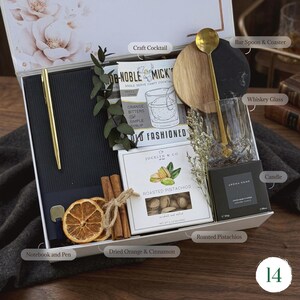 May include: A gift box with a gold pen, notebook, and a cocktail kit. Includes a whiskey glass, bar spoon, coaster, candle, and a box of roasted pistachios. Dried orange slices and cinnamon sticks add a decorative touch.