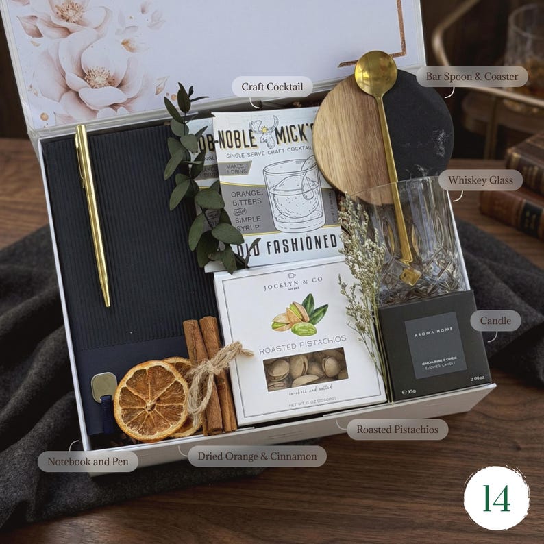 May include: A gift box with a gold pen, notebook, and a cocktail kit. Includes a whiskey glass, bar spoon, coaster, candle, and a box of roasted pistachios. Dried orange slices and cinnamon sticks add a decorative touch.