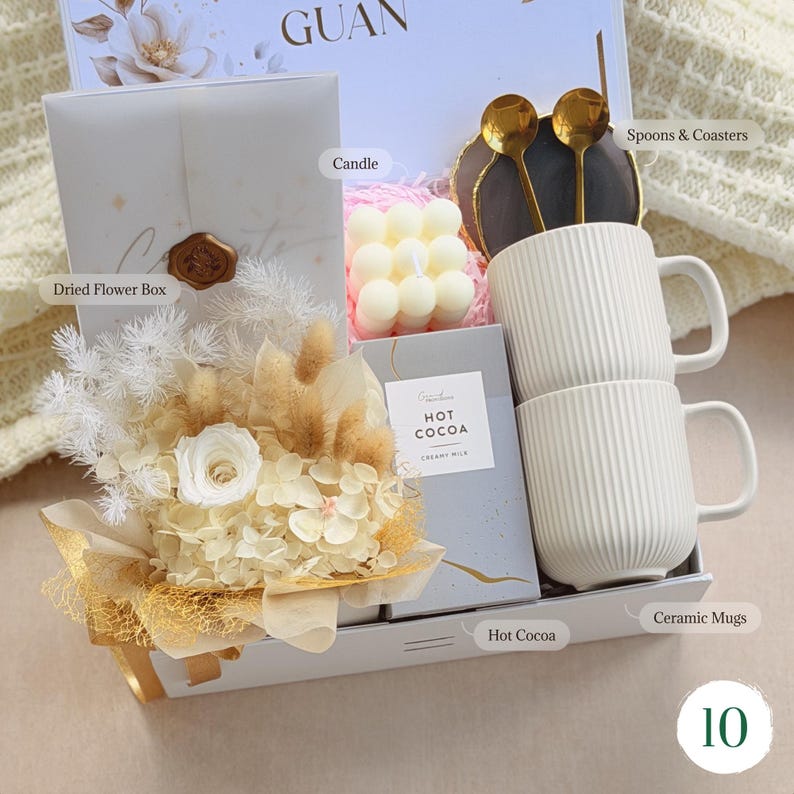 May include: A gift box containing a dried flower arrangement, two white ceramic mugs, a candle, hot cocoa, and gold spoons with coasters. The box is white with gold accents and a floral design.