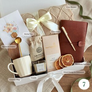 May include: A gift box containing a beige textured blanket with a white ribbon, a white ribbed ceramic mug, a gold spoon, a wooden and marble coaster, a small burlap pouch, a Moonstruck milk chocolate bar, a candle, and a brown leather notebook with a gold pen.