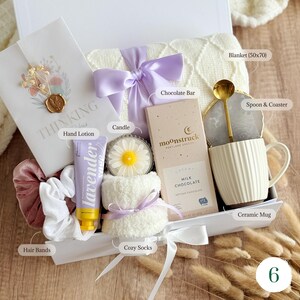 May include: A gift box filled with self-care items. Includes a white blanket (50x70 inches), a ceramic mug, a chocolate bar, a candle, hand lotion, cozy socks, hair bands, and a spoon with a coaster. The box is tied with a purple ribbon.