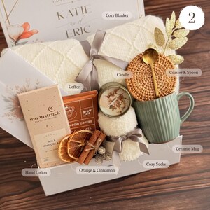 May include: A gift box with a cozy theme, featuring a cream-colored blanket, a green ceramic mug, a candle, coffee, chocolate, and cozy socks. The box also includes a coaster with a spoon, dried oranges, and cinnamon sticks. The box is tied with a gray ribbon.
