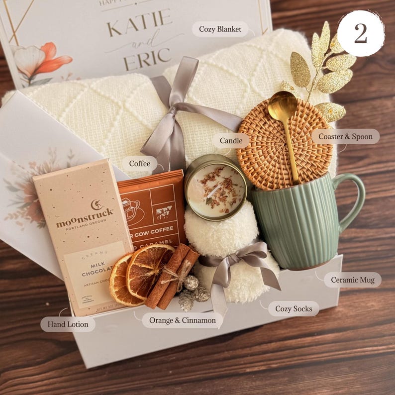 May include: A gift box with a cozy theme, featuring a cream-colored blanket, a green ceramic mug, a candle, coffee, chocolate, and cozy socks. The box also includes a coaster with a spoon, dried oranges, and cinnamon sticks. The box is tied with a gray ribbon.