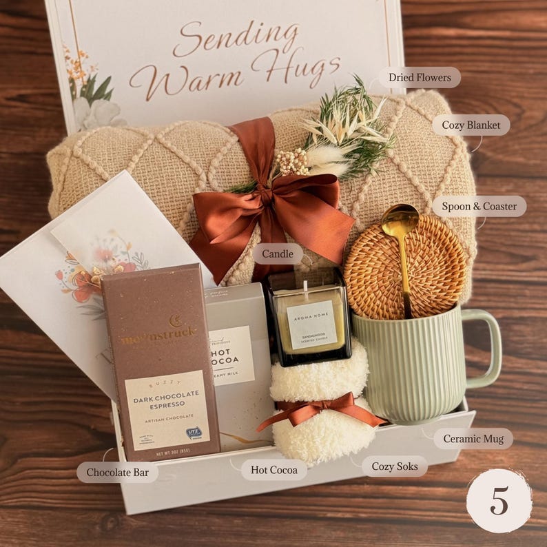 May include: A gift box labeled Sending Warm Hugs containing a beige textured blanket with a rust-colored ribbon, dried flowers, a dark chocolate espresso bar, hot cocoa mix, a candle, a pair of white fuzzy socks, a rattan coaster, and a green mug.
